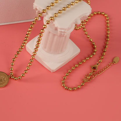 The Mia Ishaaq Beaded Chain Coin Pendant Necklace, featuring an 18k gold-plated chain, elegantly drapes over a small white column set on a pink surface.