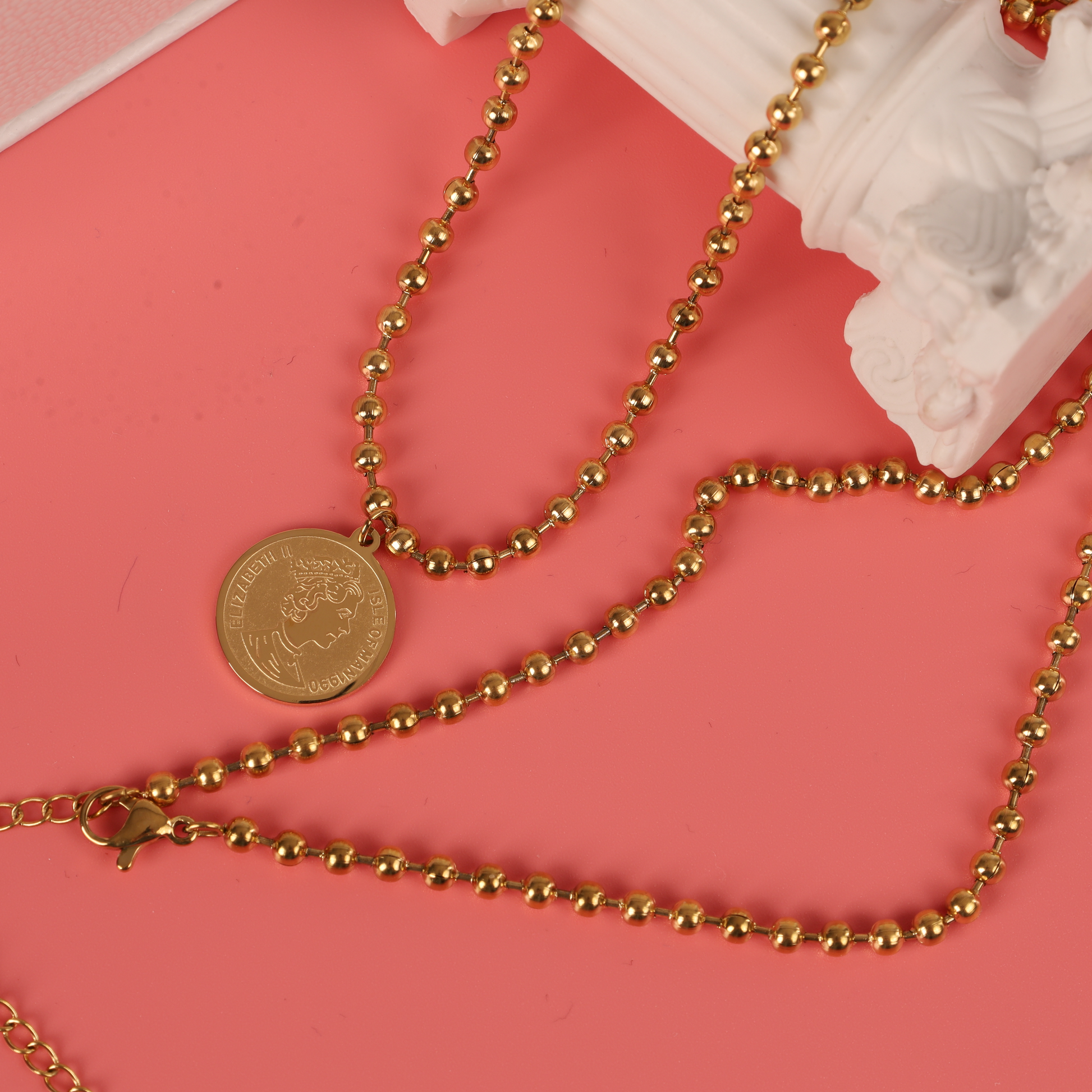 The Beaded Chain Coin Pendant Necklace by Mia Ishaaq, showcasing an exquisite 18k gold plating and a beaded chain, elegantly rests on a pink surface.