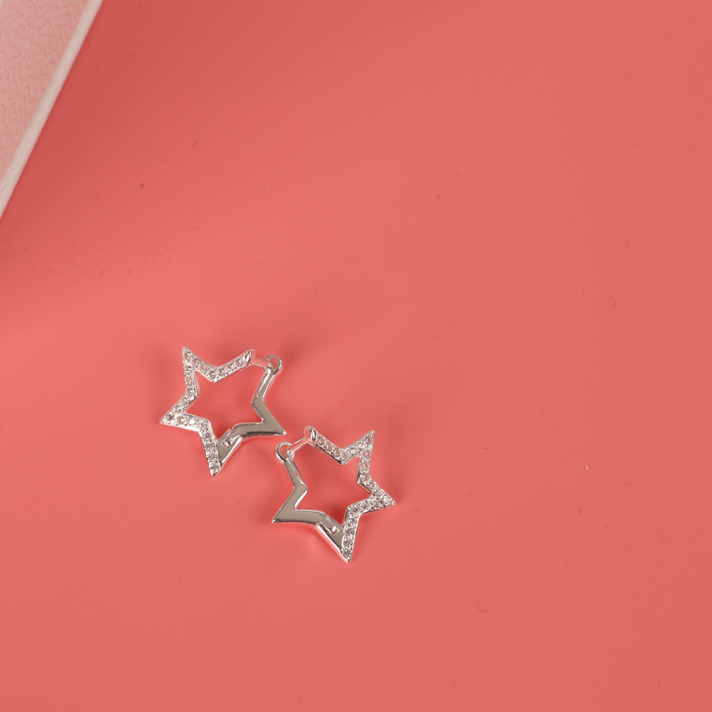 The Sparkly Star Huggie Hoop Earrings by Mia Ishaaq, crafted from 925 sterling silver, shimmer on a coral pink surface.