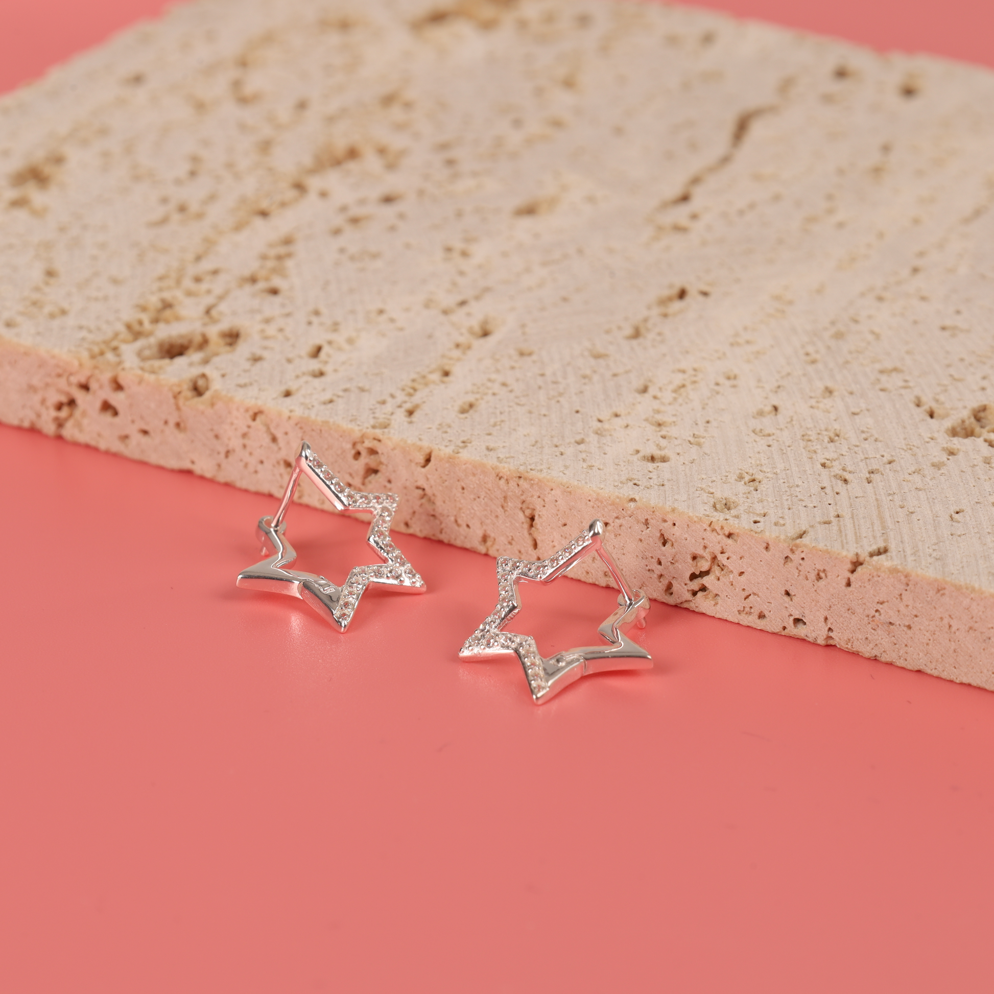 The Sparkly Star Huggie Hoop Earrings by Mia Ishaaq shimmer on a textured beige surface, set against a soft pink background.