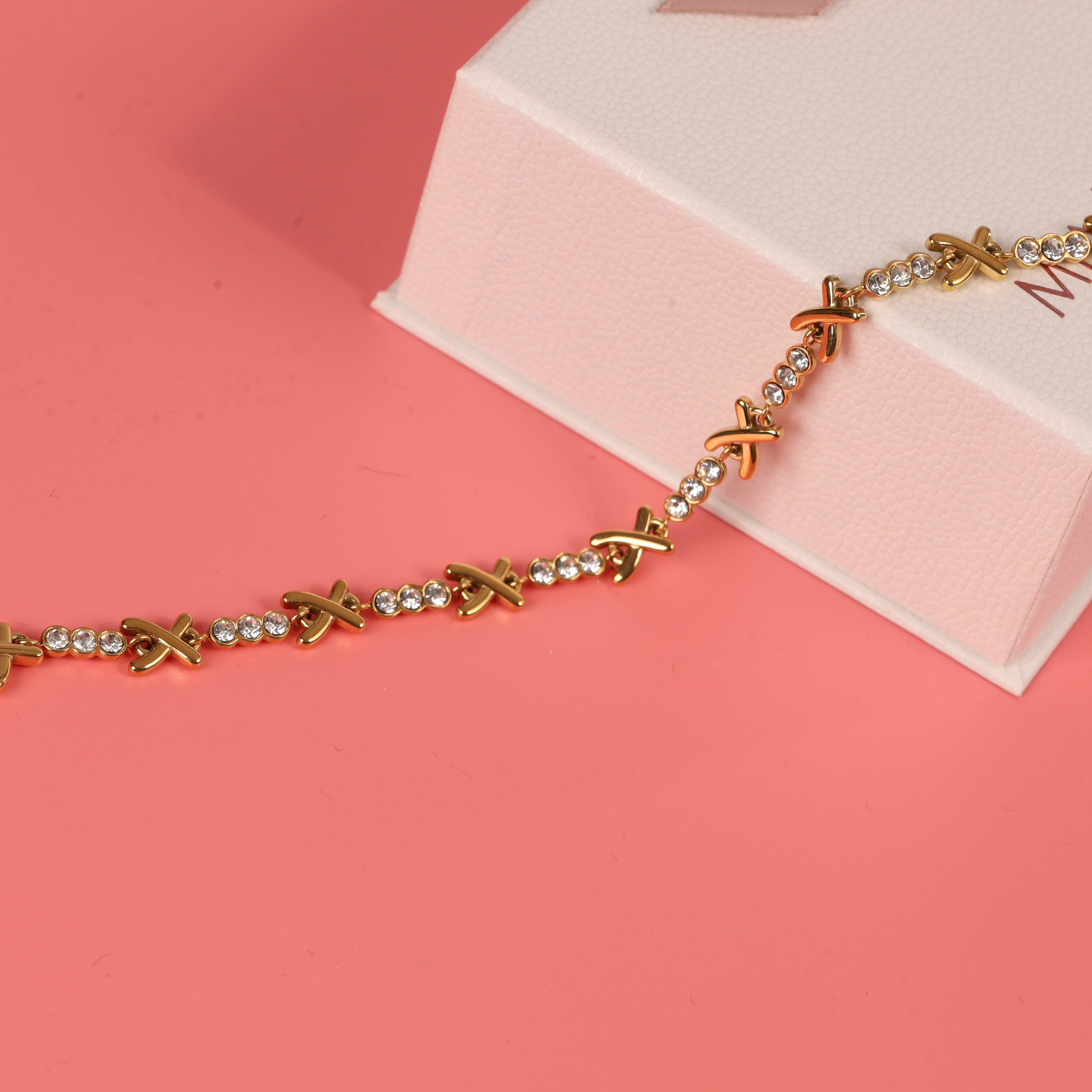 The Gemma XO Crystal Cross Bracelet - Gold by Mia Ishaaq, showcasing a refined X design, lies elegantly on a pink surface beside its stylish box.