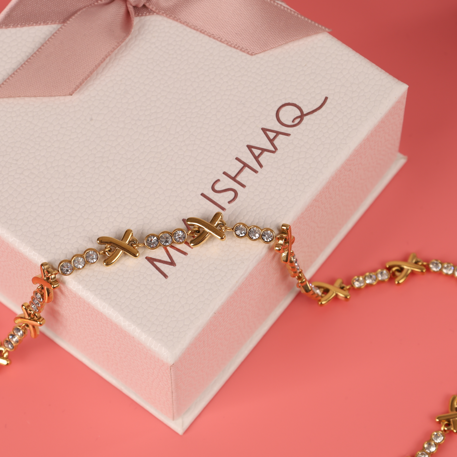 The Mia Ishaaq Gemma XO Crystal Cross Bracelet in gold elegantly complements a gold necklace with crystals, all nestled beside a white gift box adorned with a pink bow against a soft pink background.