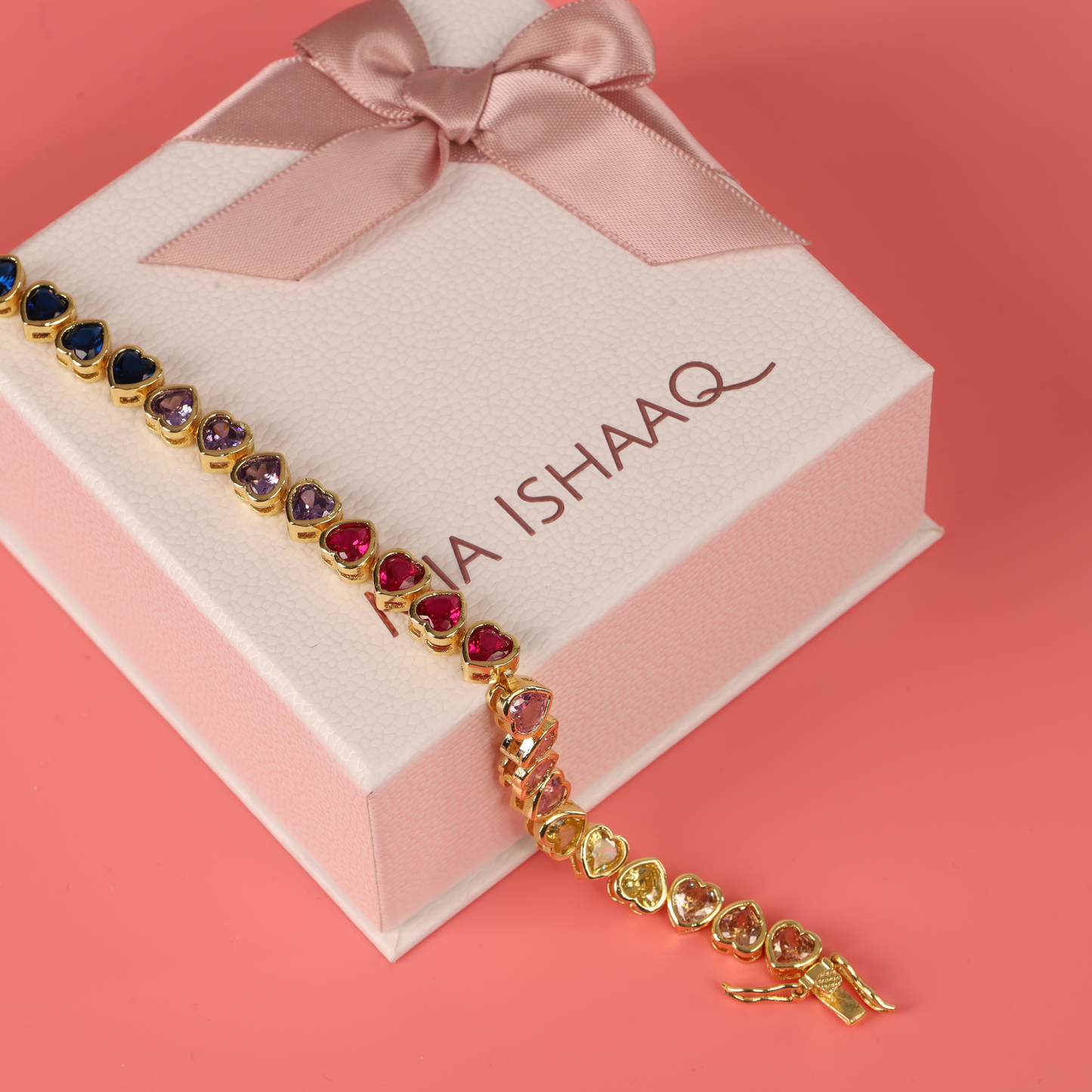 The Mia Ishaaq Rainbow Heart Tennis Bracelet, featuring an array of multicolored stones, rests elegantly on a gift box adorned with a pink ribbon, set against a coral background.