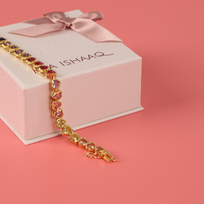 The Rainbow Heart Tennis Bracelet by Mia Ishaaq, crafted in exquisite 18K gold and adorned with multi-colored stones, is elegantly draped over a pink gift box with ribbon on a soft pink background.