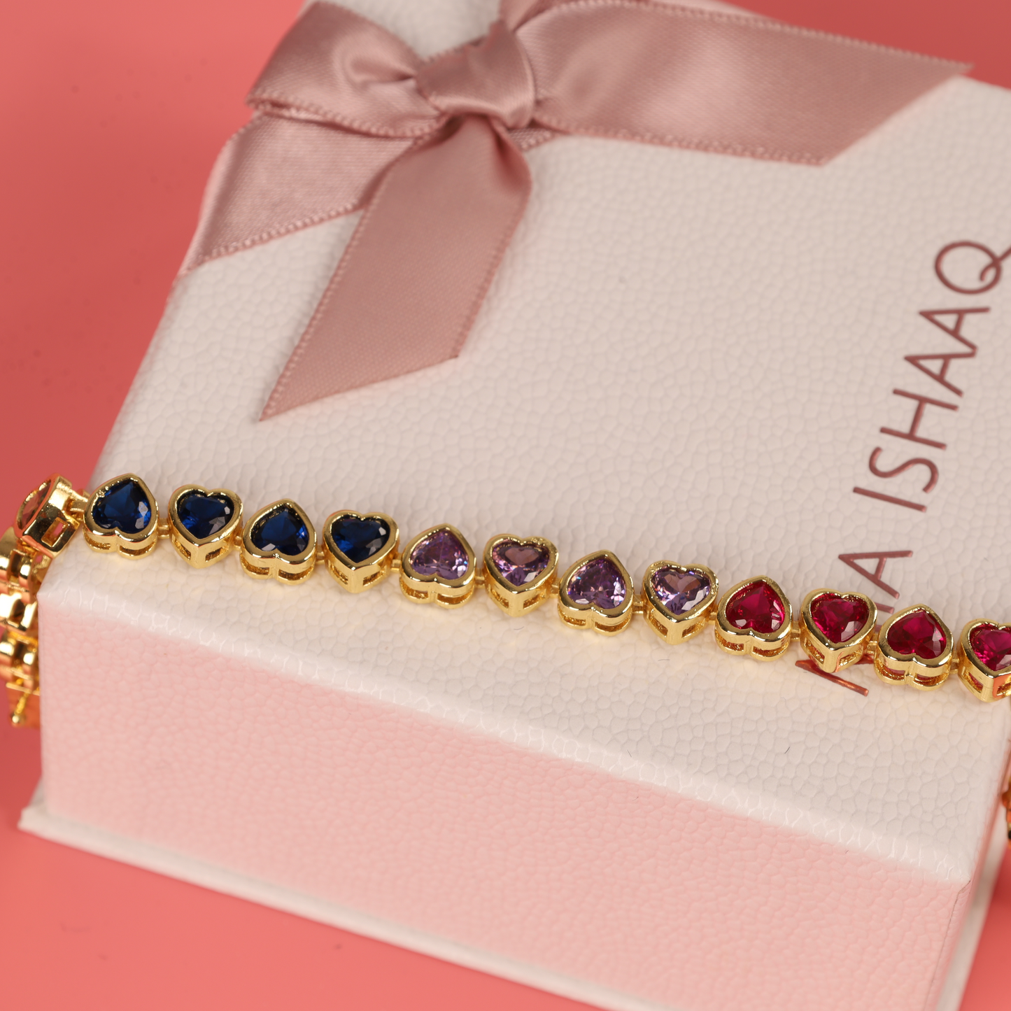 The Rainbow Heart Tennis Bracelet by Mia Ishaaq showcases heart-shaped, multicolored stones elegantly presented in a white box tied with a pink ribbon.