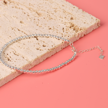 The Sterling Silver Beaded Bracelet by Mia Ishaaq, featuring a minimalist design with a small heart charm, is elegantly displayed on a textured stone surface against a coral background, highlighting its high-quality craftsmanship.