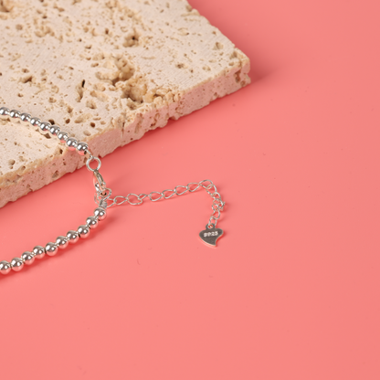 The Mia Ishaaq Sterling Silver Beaded Bracelet, featuring a heart-shaped charm, is elegantly displayed on a pink background next to a textured beige block, capturing the essence of minimalist design.