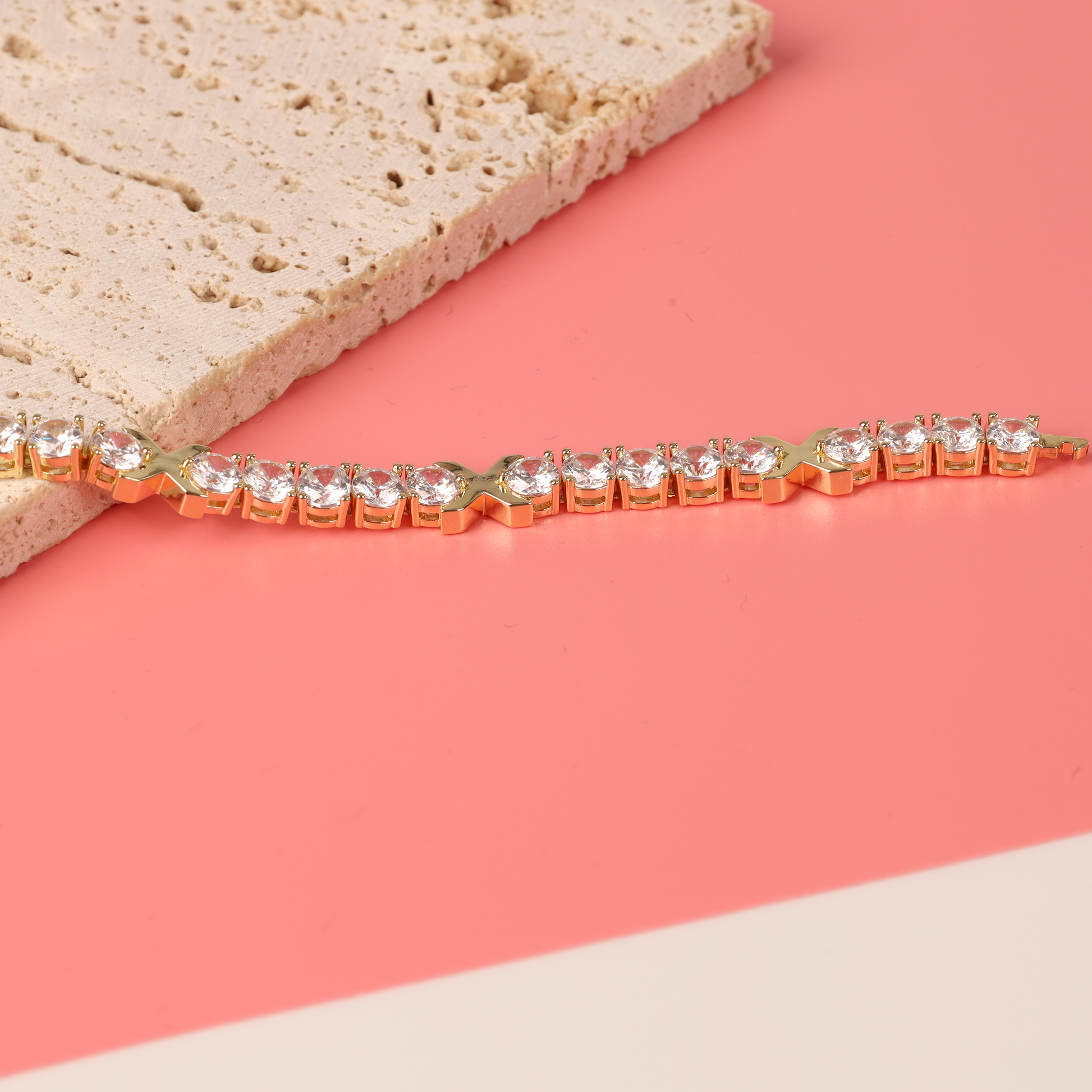 The Gemma XO Cross Love Bracelet - Gold by Mia Ishaaq, adorned with dazzling diamonds and cubic zirconia, rests elegantly on a pink surface against a stone background.