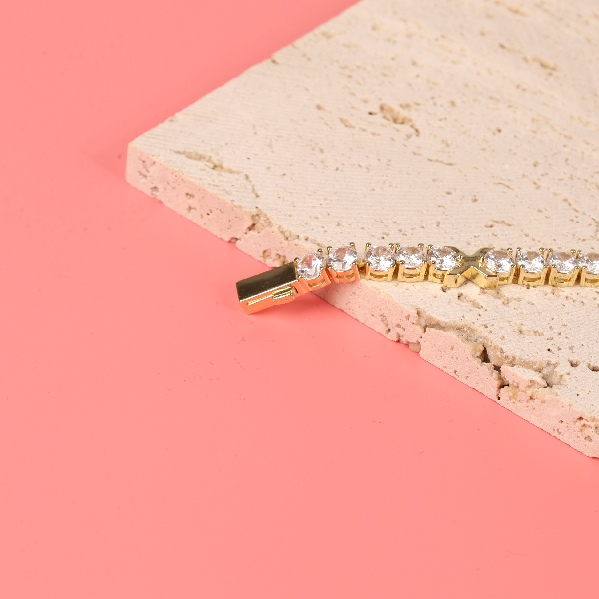 A stunning Gemma XO Cross Love Bracelet - Gold by Mia Ishaaq sparkles elegantly with cubic zirconia on a textured stone slab against a coral background.