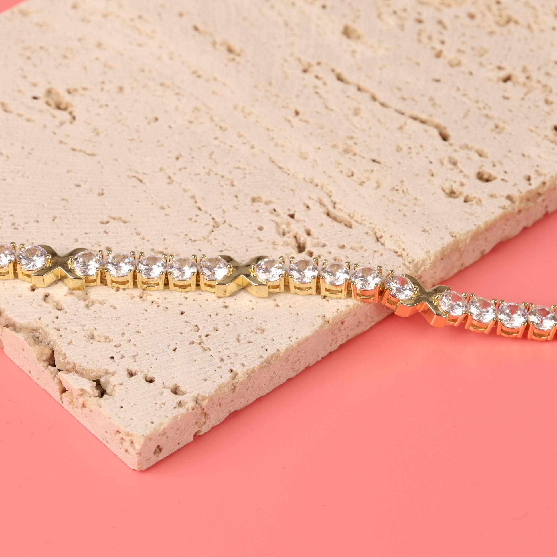 The Gemma XO Cross Love Bracelet in gold by Mia Ishaaq rests elegantly on a textured stone surface against a pink background.