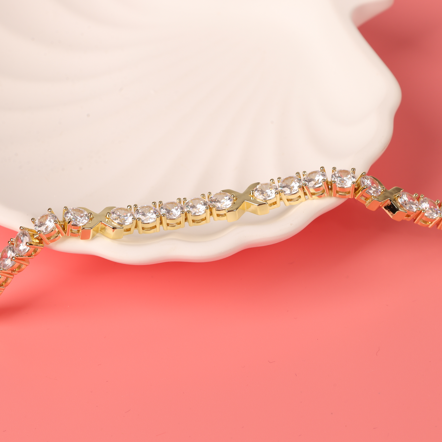 The Gemma XO Cross Love Bracelet - Gold from Mia Ishaaq, adorned with dazzling diamond-like stones, elegantly rests on a pink surface near a white seashell. Its shimmering gold plating enhances the charming allure of this exquisite piece.