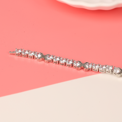 The chic Gemma XO Cross Love Bracelet in silver by Mia Ishaaq, adorned with sparkling gemstones, shines against a pink and white background. Its elegant design, inspired by Gemma Owen, is enhanced with 18k white gold plating for a luxurious touch.