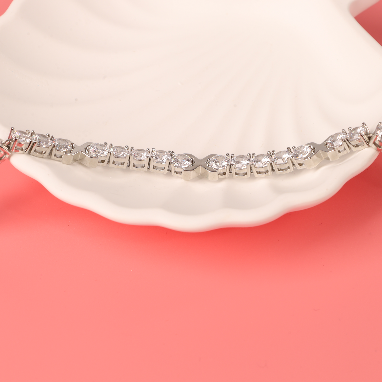 The Gemma XO Cross Love Bracelet - Silver by Mia Ishaaq, adorned with sparkling stones and 18k white gold plating, rests elegantly on a white seashell against a pink background.