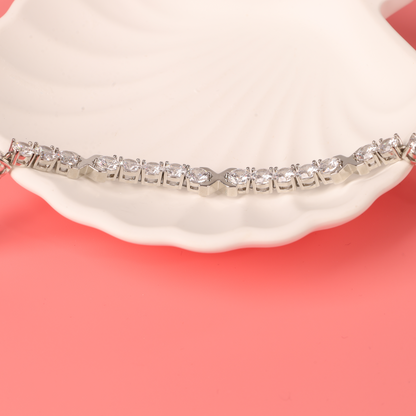 The Gemma XO Cross Love Bracelet - Silver by Mia Ishaaq, adorned with sparkling stones and 18k white gold plating, rests elegantly on a white seashell against a pink background.