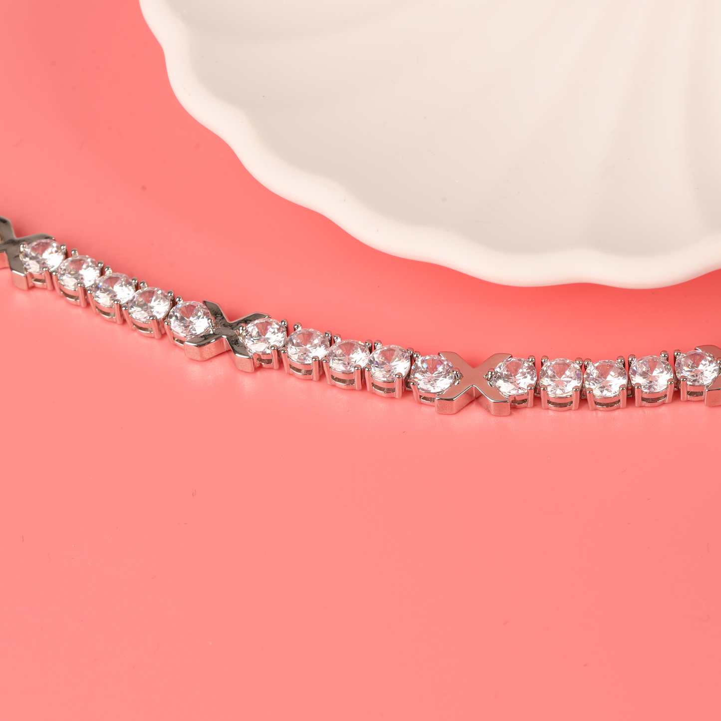 The Gemma XO Cross Love Bracelet - Silver from Mia Ishaaq, with its clear gemstones and 18k white gold plating, sparkles elegantly on a coral pink surface beside a white ceramic dish.