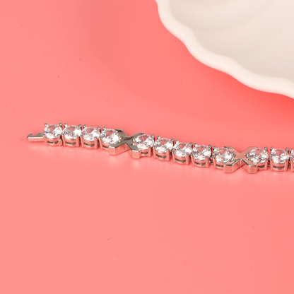 Gemma XO Cross Love Bracelet - Silver by Mia Ishaaq, featuring sparkling gemstones and 18k white gold plating, elegantly displayed on a coral pink surface next to a white scalloped-edge dish.