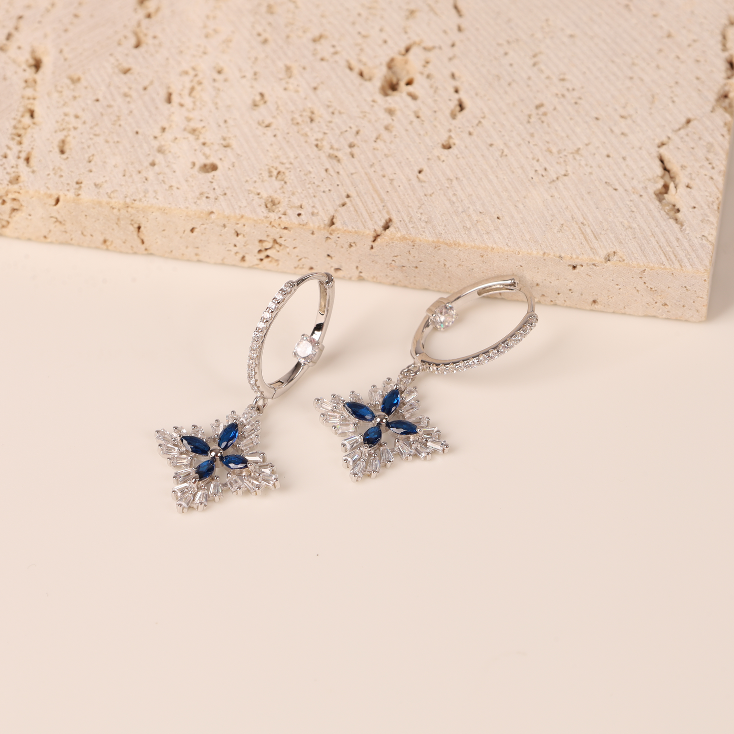 The Mia Ishaaq Blue Spinel CZ Earrings, crafted from 925 sterling silver and featuring dazzling blue spinel cubic zirconia, elegantly rest on a lightly textured surface.