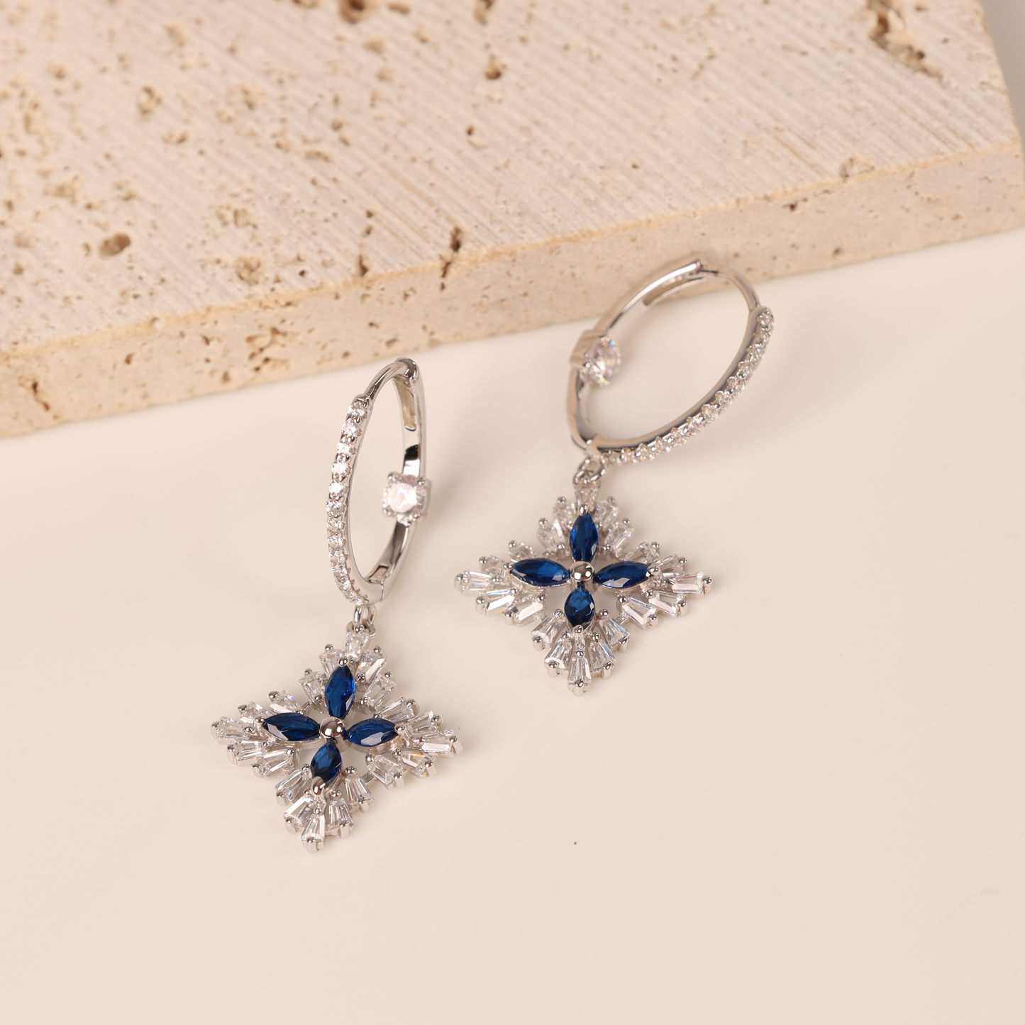 Mia Ishaaq's Blue Spinel CZ Earrings, crafted from 925 Sterling Silver, dazzle with clear gemstones against a textured beige backdrop.