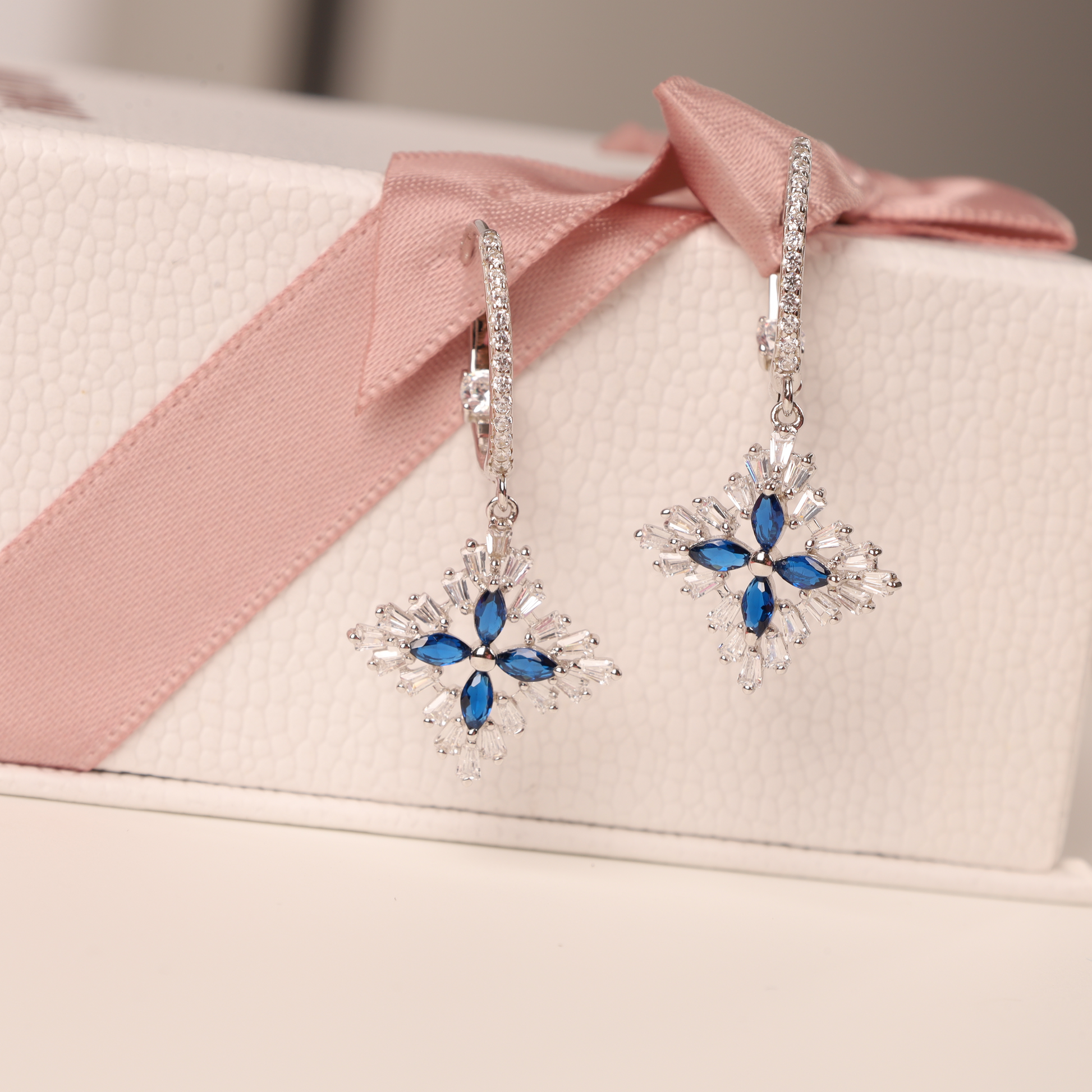 The Blue Spinel CZ Earrings by Mia Ishaaq, crafted from 925 sterling silver, rest elegantly on a white box with a pink ribbon.