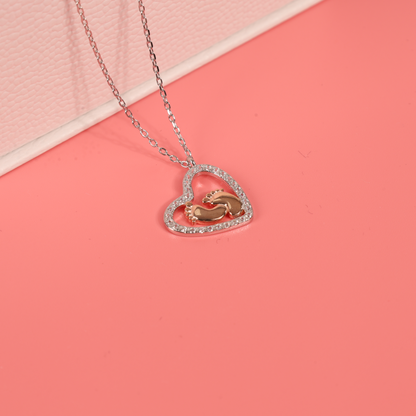 Close up image of a Sterling Silver Cubic Zirconia Mothers Love Heart Pendant with baby footprints necklace on a pink background - Mia Ishaaq