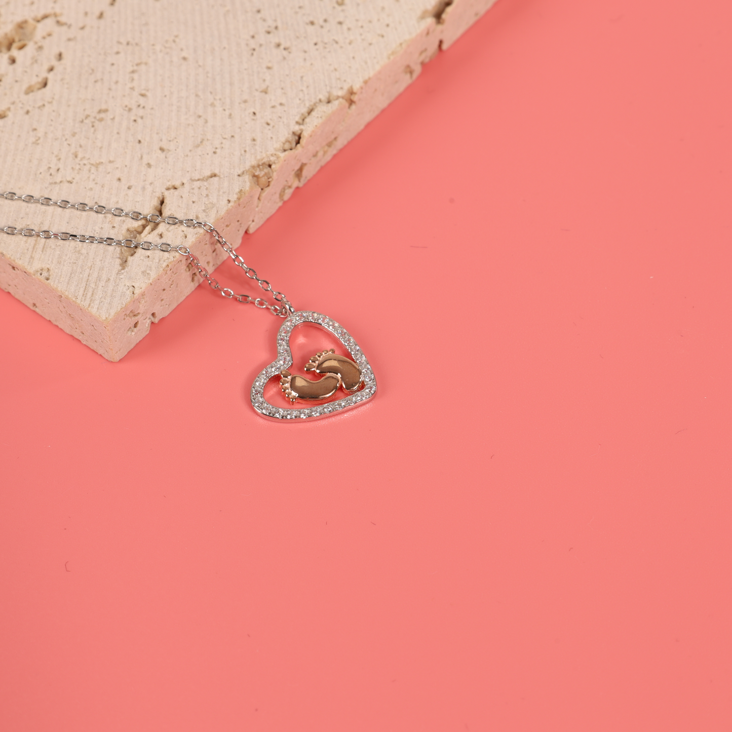 Detail close up of the Sterling Silver Cubic Zirconia Mothers Love Heart Pendant with baby footprints necklace placed on a natural colour prop with a pink background - Mia Ishaaq