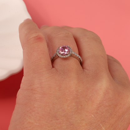 Model holding the ring delicately between their fingers, allowing a clear view of the pink cubic zirconia and surrounding halo stones - Mia Ishaaq