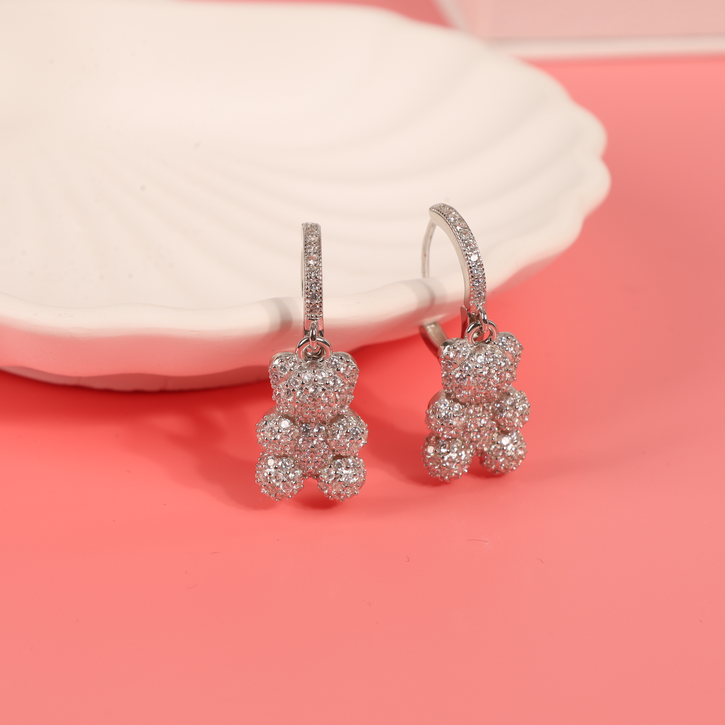 A stunning display of sterling silver teddy bear dangle earrings on a pink background, presented on a white ceramic shell plate for an eye-catching aesthetic - Mia Ishaaq