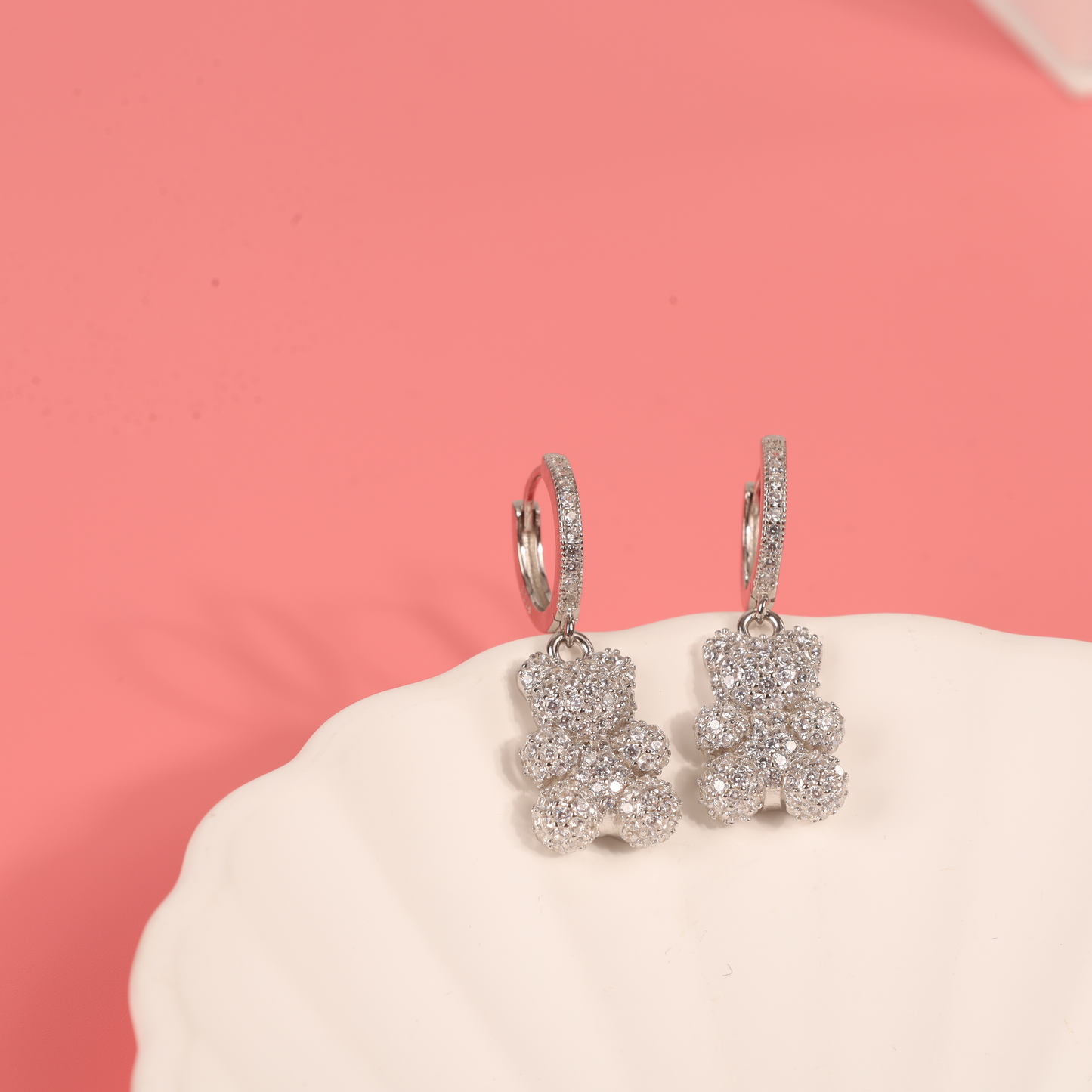 Overhead shot of teddy bear dangle earrings resting on a pink background with a white ceramic shell plate, capturing the intricate cubic zirconia details and nostalgic charm - Mia Ishaaq