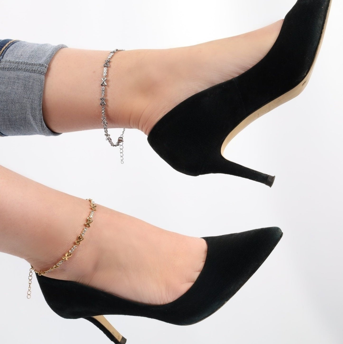 Two legs adorned in black high heels and stylish ankle bracelets—one featuring the Gemma XO Crystal Cross Anklet by Mia Ishaaq, gold-plated with a crystal cross, and the other with an adjustable fit anklet—stand elegantly against a white background.