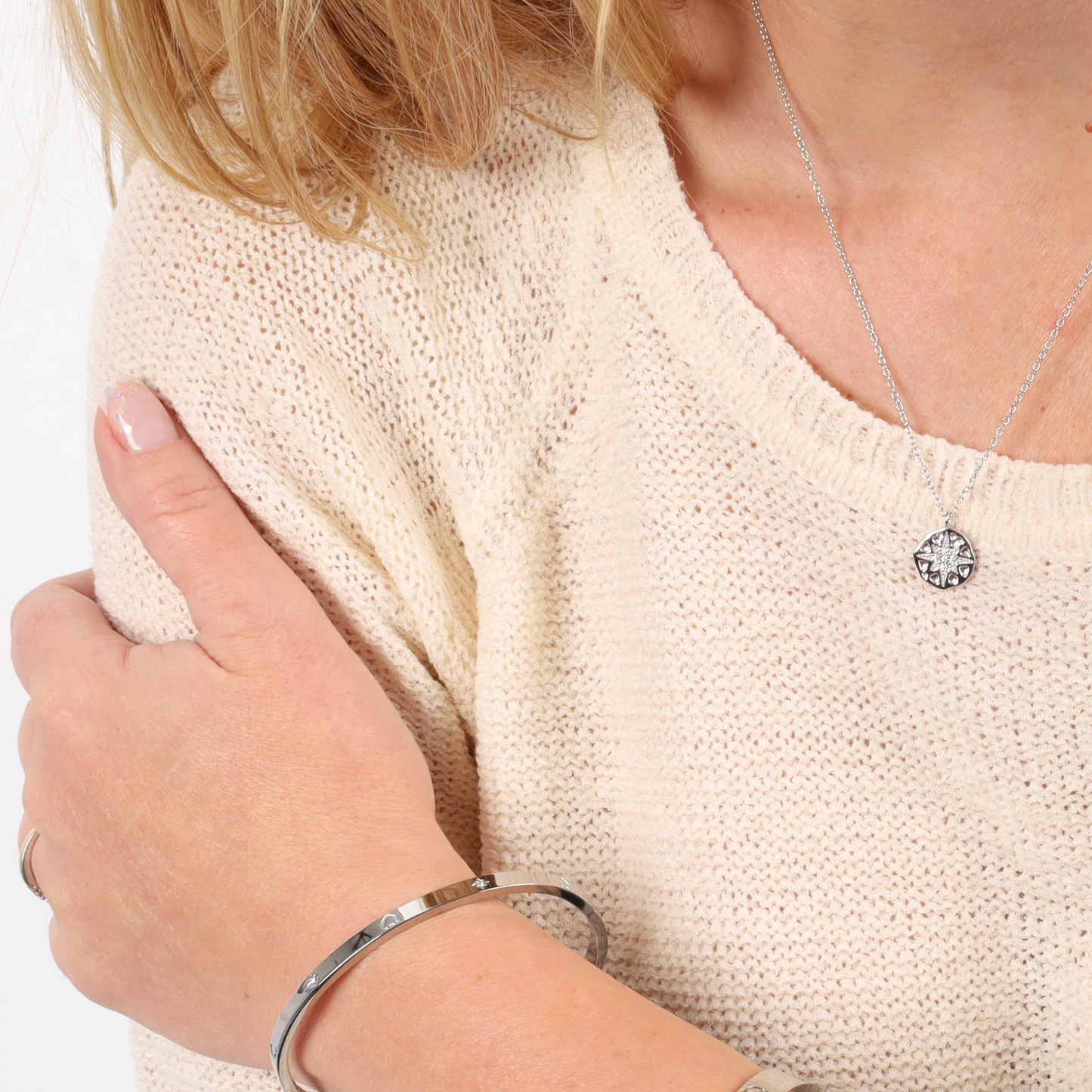 A person, adorned with a beige sweater, holds their arm, showcasing a bracelet and the stunning Polaris Coin Pendant Necklace - Silver from Mia Ishaaq®, featuring a North Star pendant.