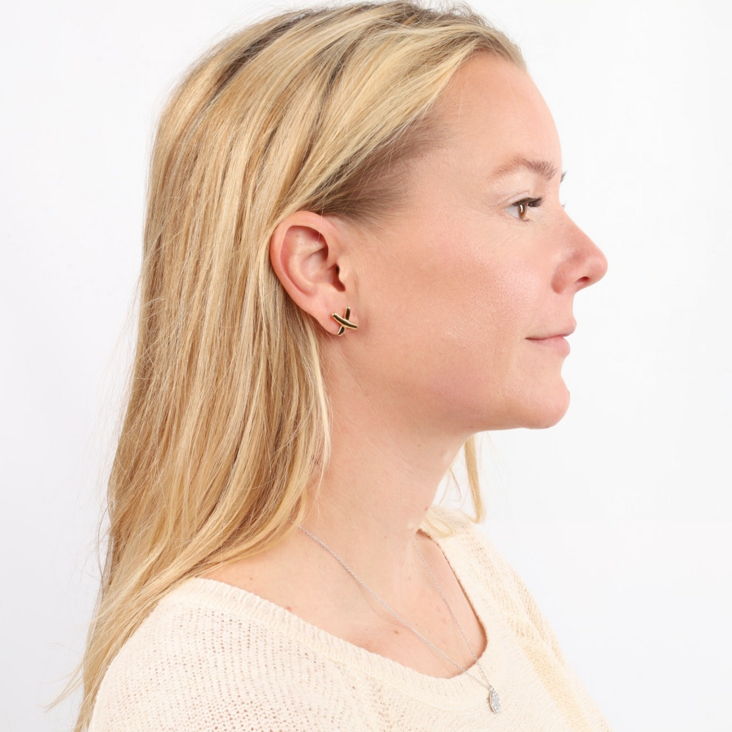 A woman with long blonde hair, elegantly adorned in a beige sweater and Mia Ishaaq® Gemma Owen Inspired XO Cross Stud Earrings - Gold featuring 18k gold plating, set against a white background.