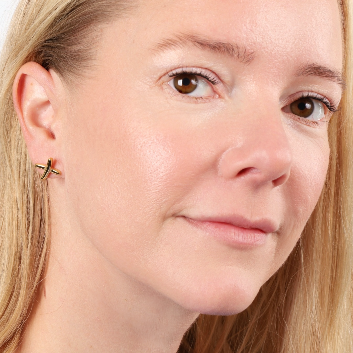 Close-up of a person with blonde hair wearing the Mia Ishaaq® Gemma Owen Inspired XO Cross Stud Earrings in gold, smiling slightly.