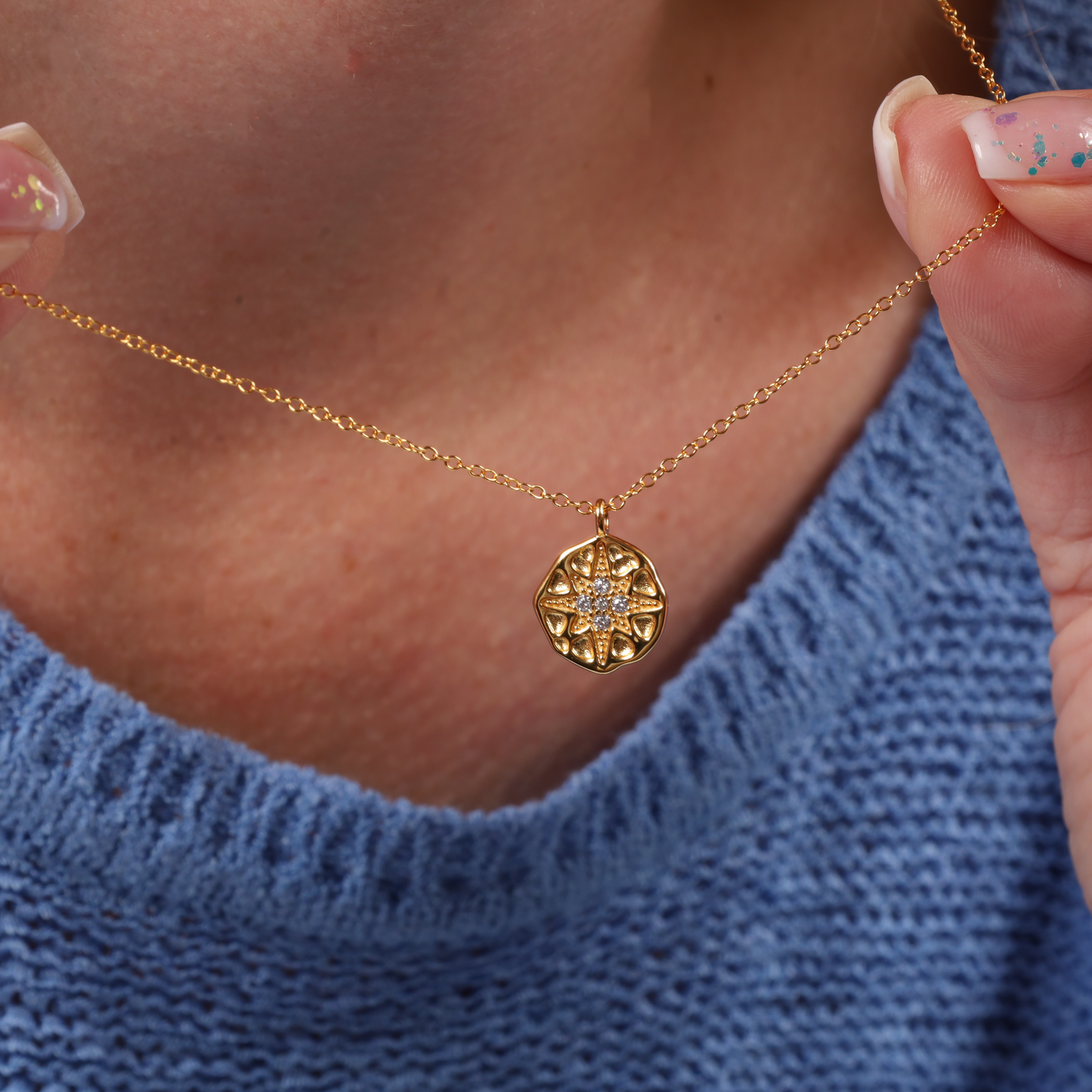 A person in a blue sweater holds the Polaris Coin Pendant Necklace - Gold by Mia Ishaaq®, featuring a gold circular pendant embellished with a star pattern and shimmering gemstones.