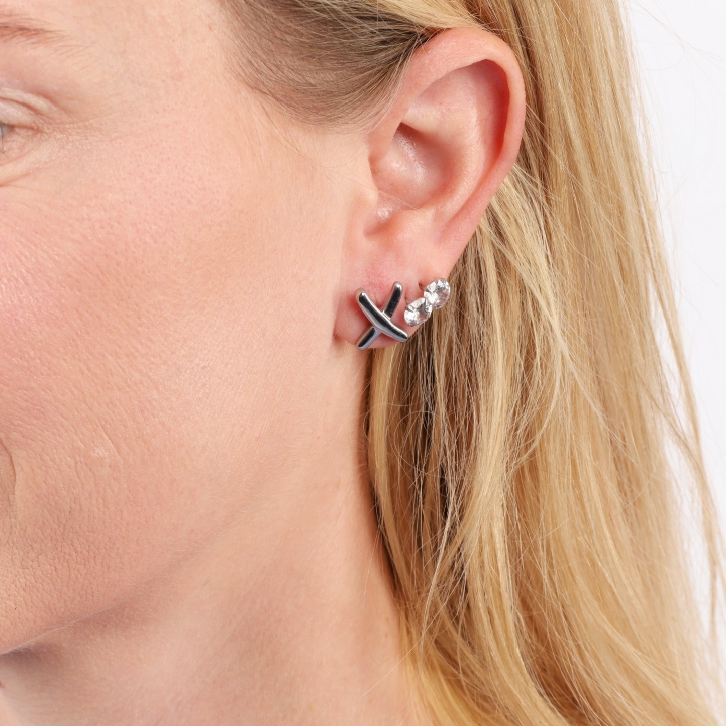 A close-up of a woman's ear adorned with minimalist stud earrings showcases the XO Cross Stud Earrings in silver by Mia Ishaaq®, featuring a black cross design reminiscent of Gemma Owen-inspired elegance, and glistening with sparkling stones.