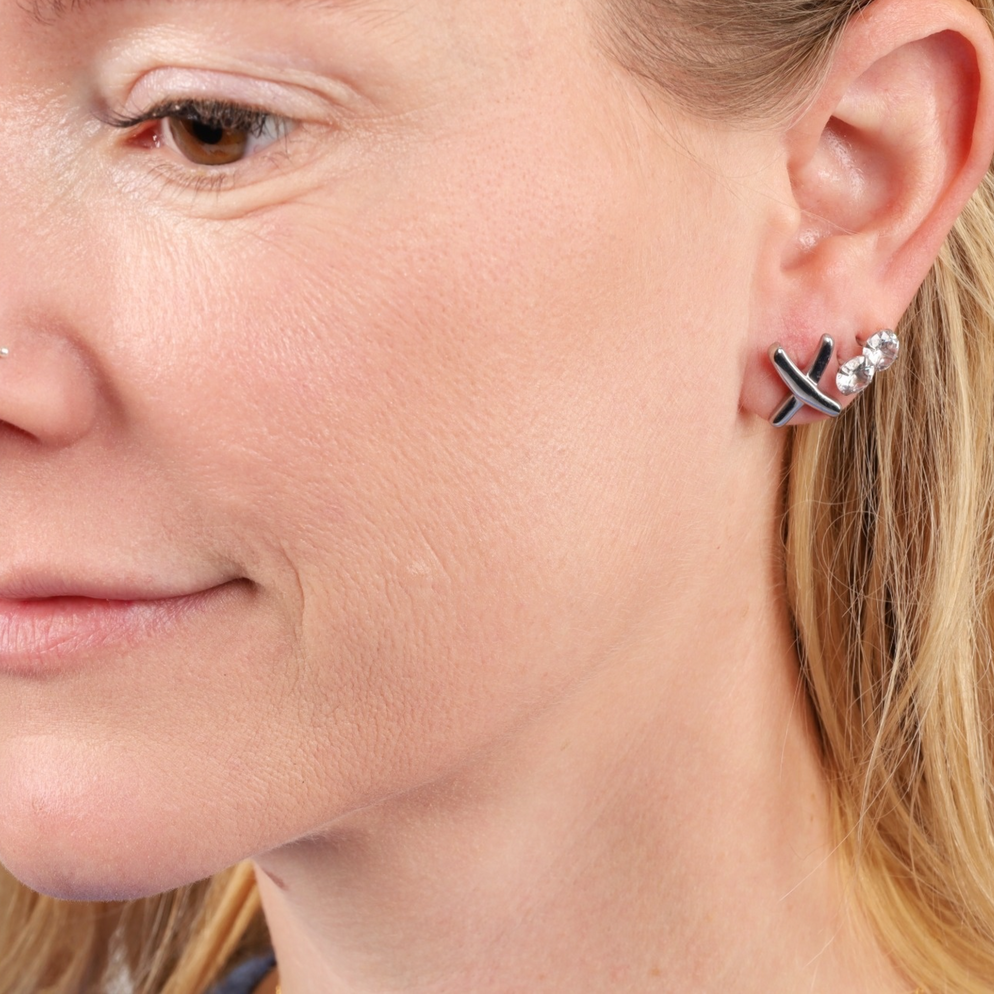 Close-up of a woman's ear adorned with Mia Ishaaq® Gemma Owen Inspired XO Cross Stud Earrings in silver, along with a row of minimalist stud earrings, creating a refined and stylish look.