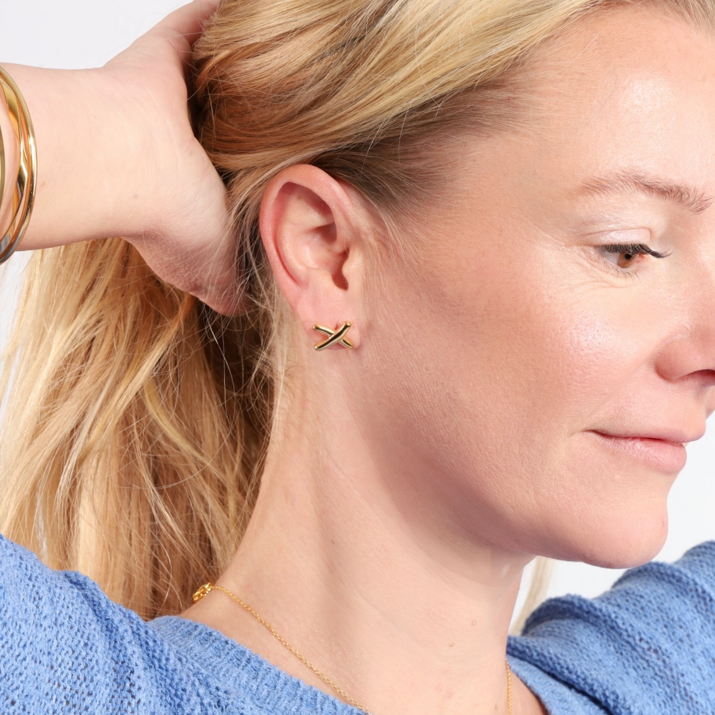 A woman with long blonde hair wears Gemma Owen Inspired XO Cross Stud Earrings in gold by Mia Ishaaq®, paired with a blue top, her expression relaxed as she gazes to the side.