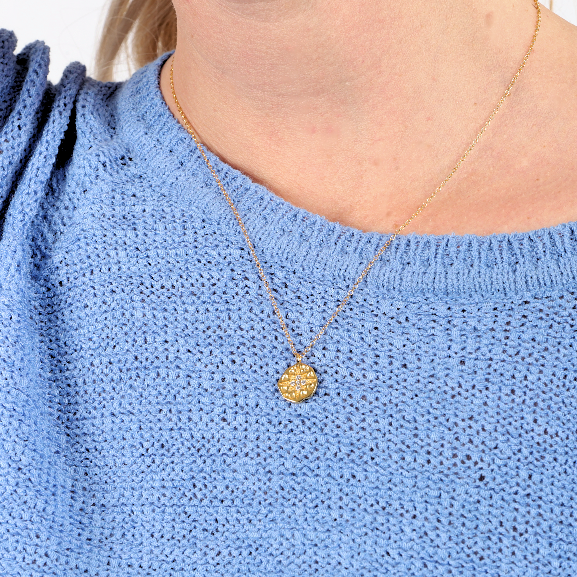 A person wearing the Polaris Coin Pendant Necklace - Gold by Mia Ishaaq® on a thin chain, perfectly complemented by a blue knit sweater.