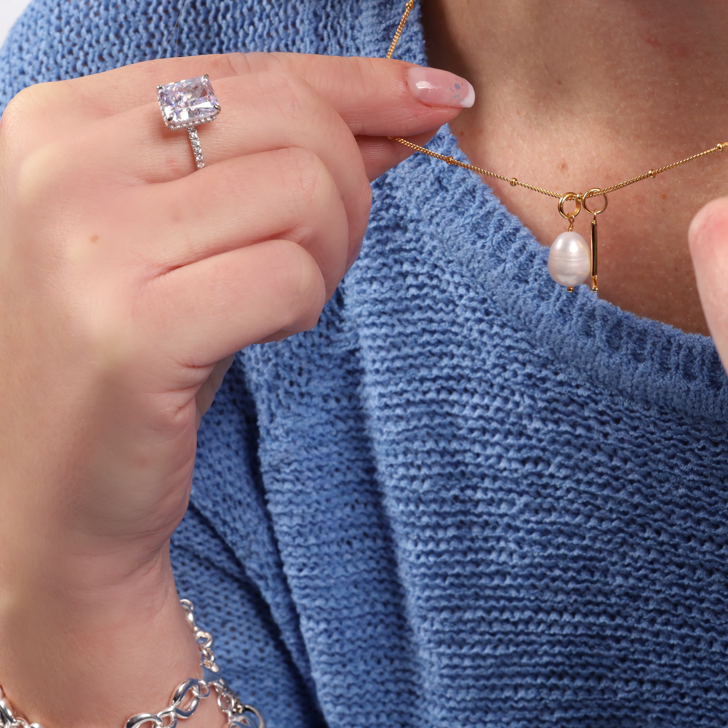 A person in a blue sweater is holding a necklace with a pearl pendant while wearing the stunning Crushed Ice Lilac Radiant Promise Ring by Mia Ishaaq and an elegant bracelet.