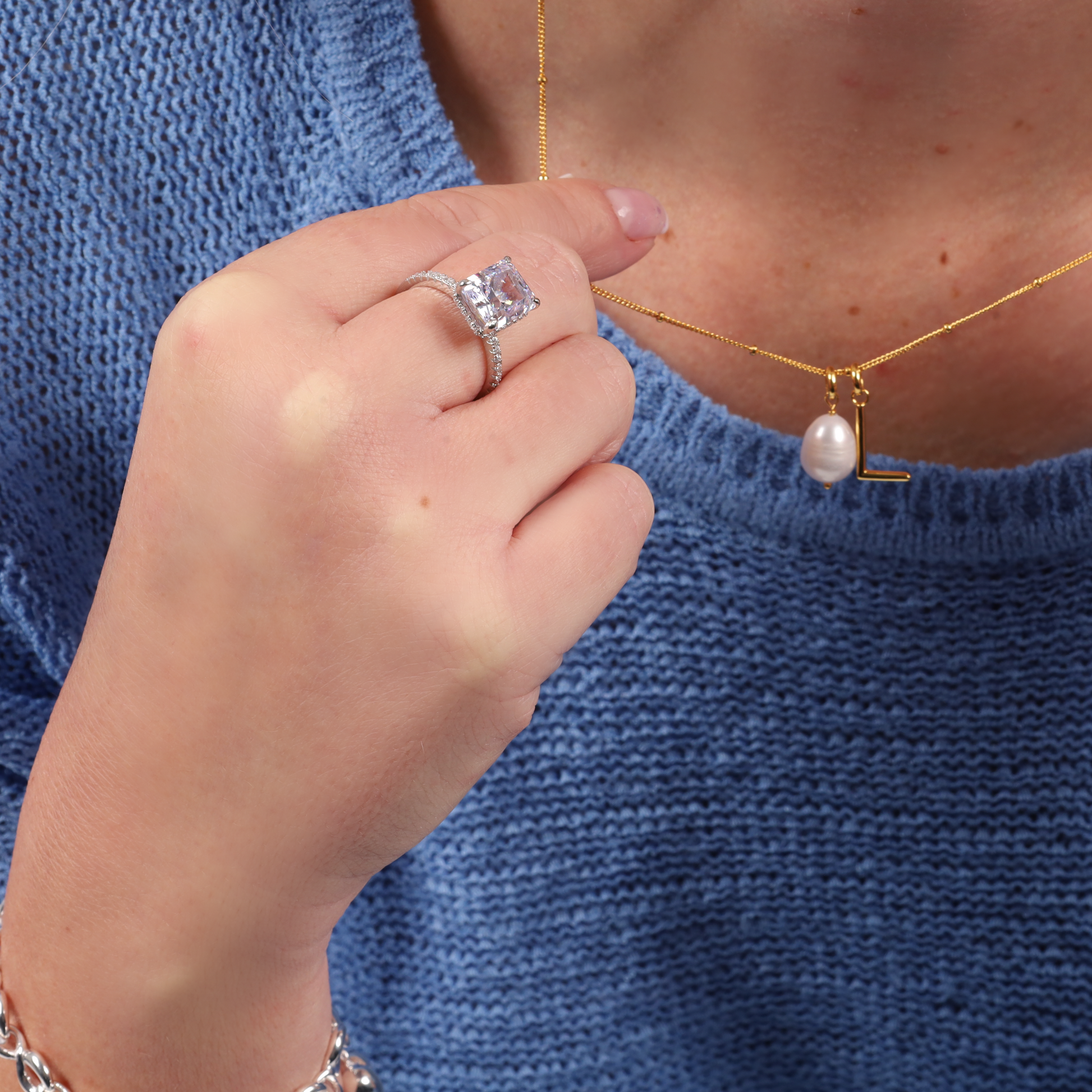 A person in a blue sweater elegantly displays a pearl necklace and the stunning Crushed Ice Lilac Radiant Promise Ring by Mia Ishaaq on their hand.