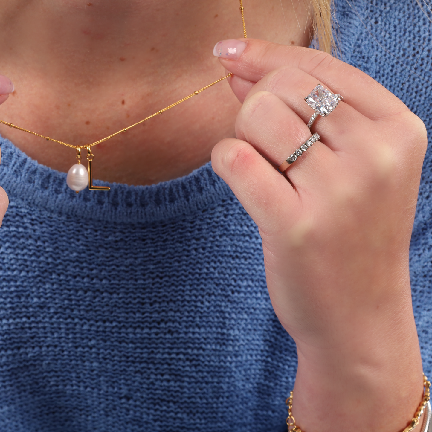 A person wearing a blue sweater, holding a pearl necklace, showcases the Sterling Silver Clear Radiant Ring by Mia Ishaaq on their finger.