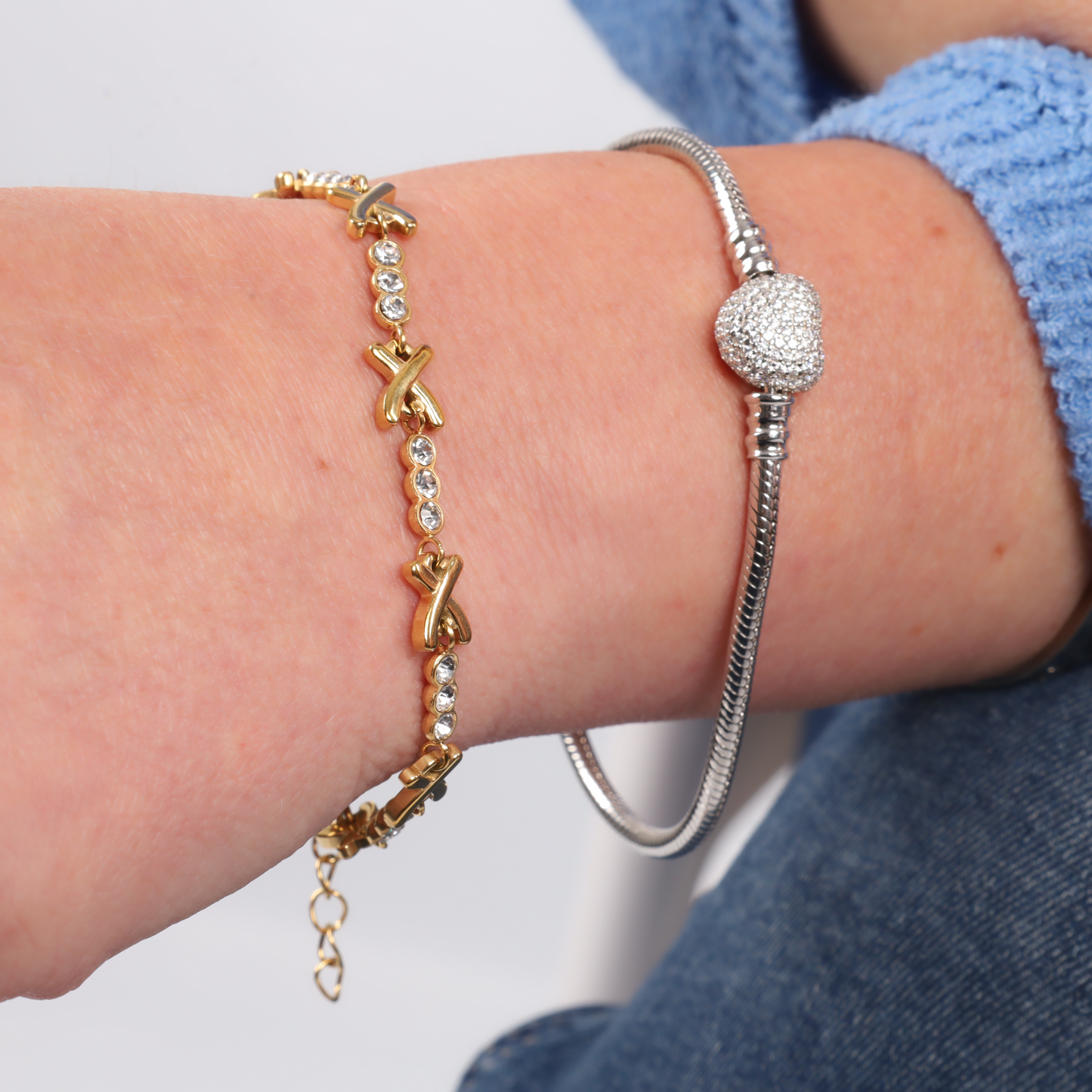 A close-up of a wrist adorned with the Gemma XO Crystal Cross Bracelet - Gold by Mia Ishaaq, featuring X-shaped gold links, paired with a silver bracelet showcasing a round, sparkly charm. This waterproof hypoallergenic bracelet set effortlessly complements any style.