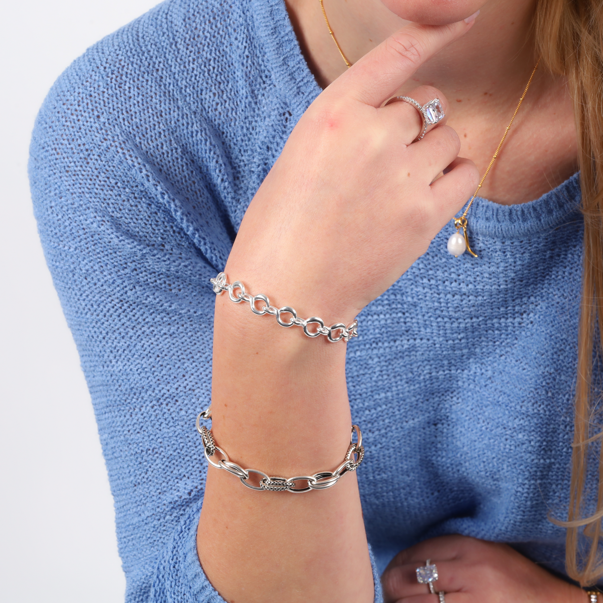 A person in a blue sweater is wearing the "Romantic Heart" Twist Chain Bracelet by Mia Ishaaq, along with a sterling silver ring and a necklace featuring a pearl pendant.