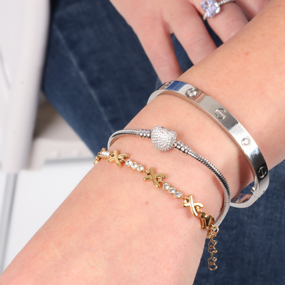 Close-up of a wrist adorned with a silver bangle, the ATHENAIE Pave Heart Snake Chain Charm Bracelet crafted from 925 sterling silver, and a gold bracelet with crystal accents and sparkling beads.