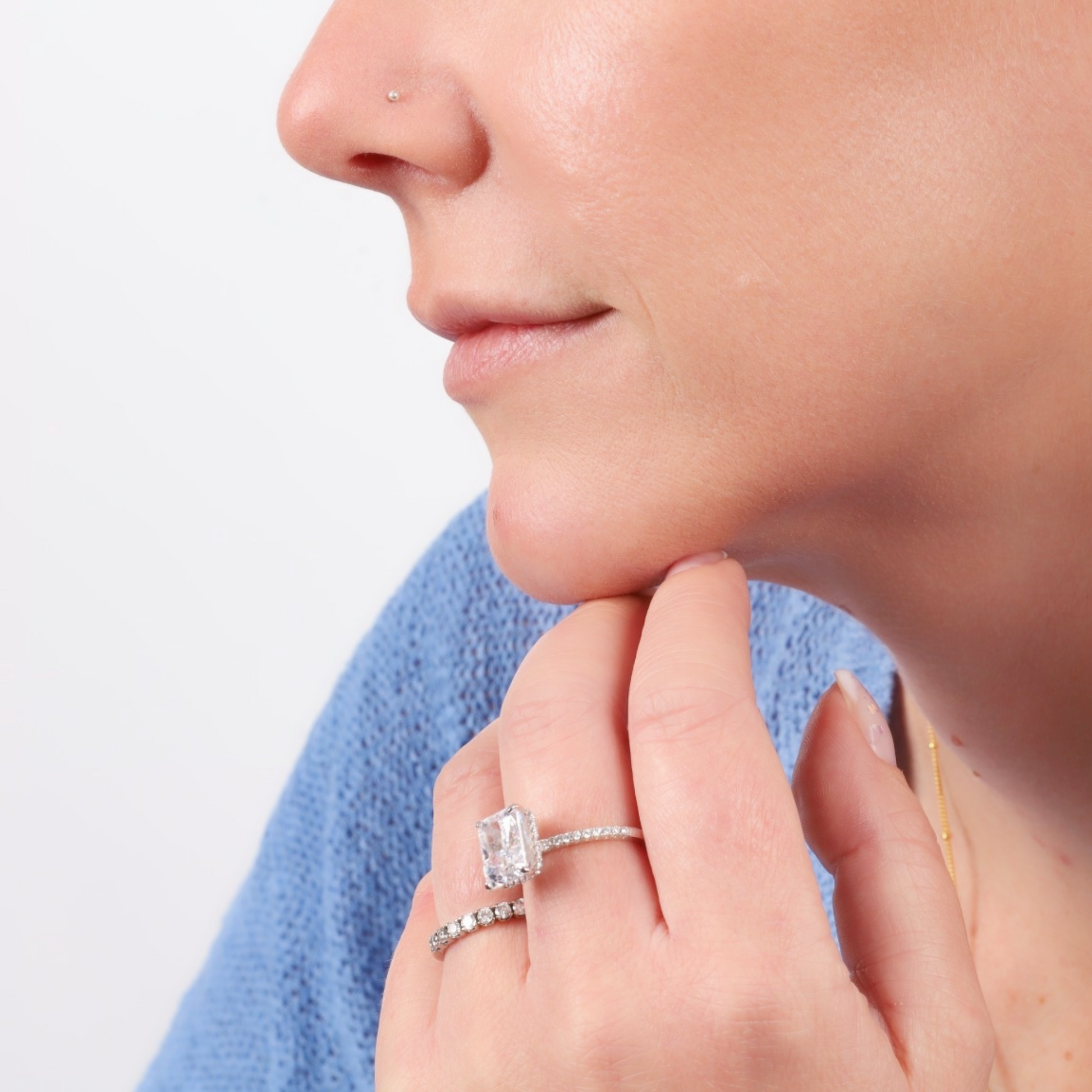 A person wearing a blue top shows off a nose piercing and two Mia Ishaaq Sterling Silver Clear Radiant Rings on their finger against a plain background.