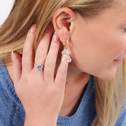 A woman with long blonde hair wearing a blue sweater showcases Shimmering Teddy Bear Earrings by Mia Ishaaq and a ring on her hand, both crafted from 925 sterling silver and adorned with dazzling cubic zirconia accents.