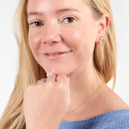 A smiling woman with long blonde hair, wearing Mia Ishaaq Gold Galaxy Huggie Hoop Earrings and rings, holds her hand near her face.