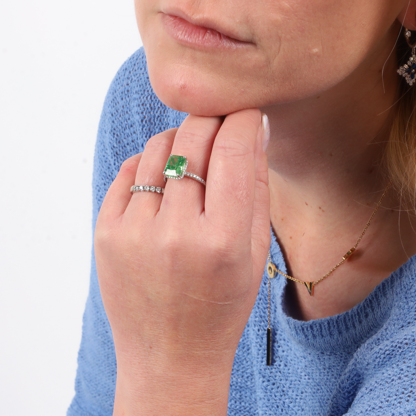 A person in a blue sweater showcases their hand, adorned with a Crushed Ice Green Radiant Promise Ring by Mia Ishaaq and a sterling silver ring, resting thoughtfully on their chin.