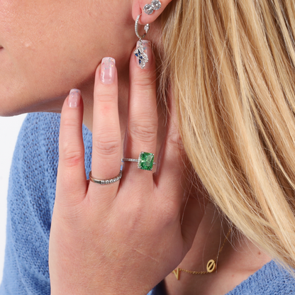 A woman wearing the Mia Ishaaq Crushed Ice Green Radiant Promise Ring and silver earrings, featuring a captivating green cubic zirconia stone, touches her face with manicured nails while donning a blue sweater.