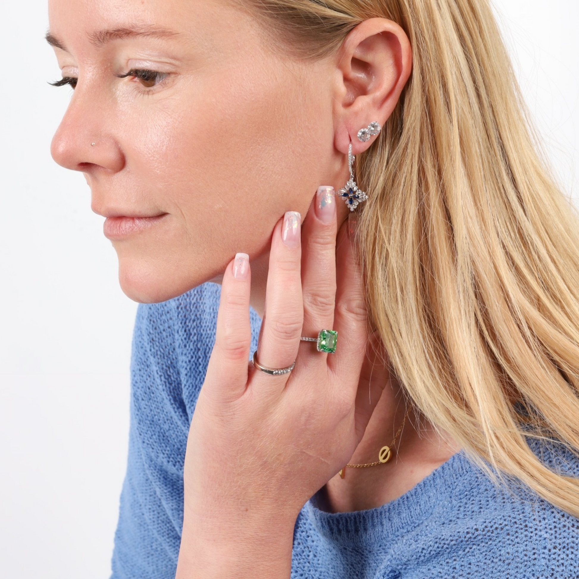 A woman with long blonde hair gazes thoughtfully, her fingers grazing her face. She's adorned with earrings and a gleaming Crushed Ice Green Radiant Promise Ring by Mia Ishaaq, featuring a green cubic zirconia stone that adds a touch of elegance to her contemplative pose.