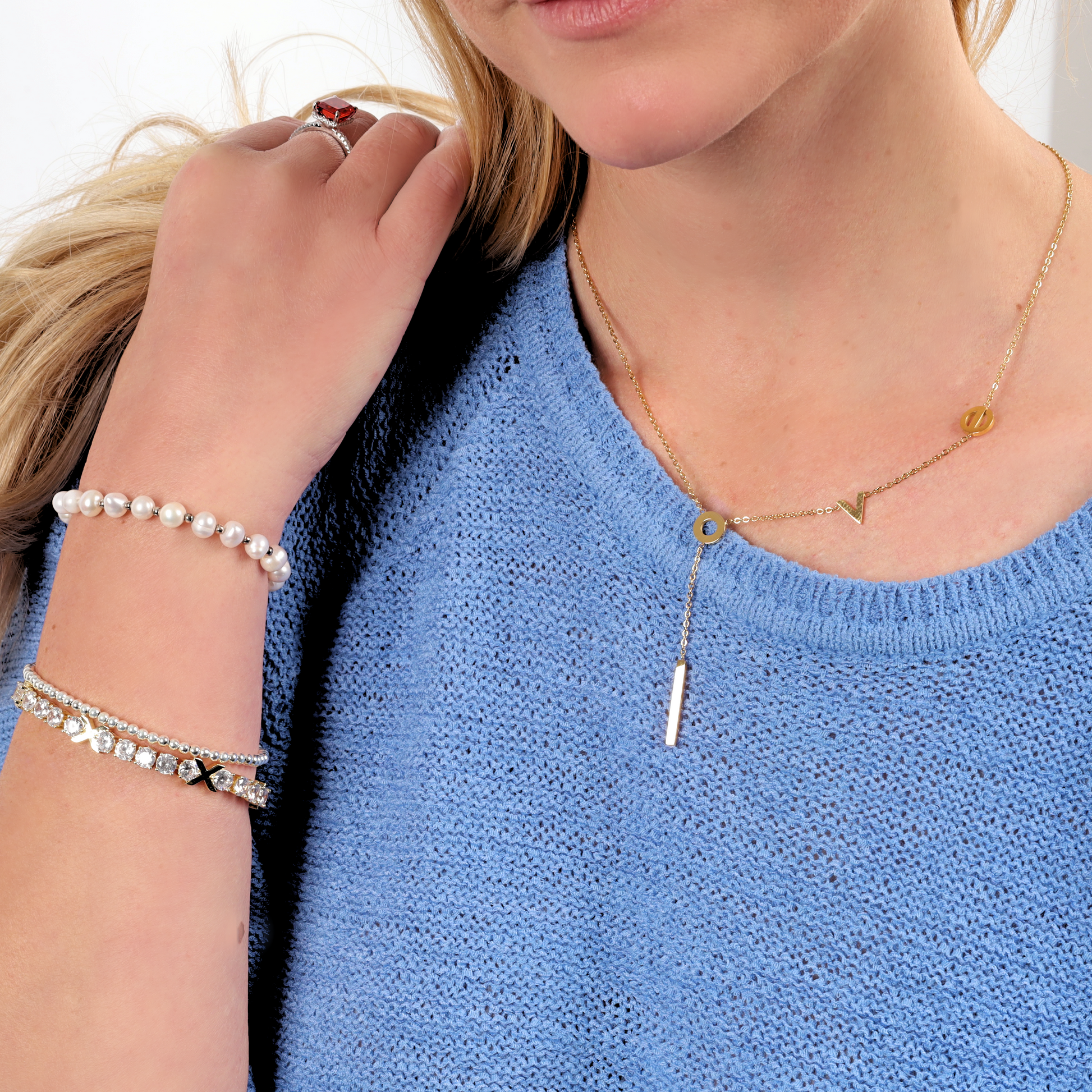 A close-up iof a person wearing a water-resistant gold necklace with the letters "L", "O", "V", and "E" arranged along the chain, ending in a vertical bar pendant. The person is dressed in a light blue knit sweater and accessorized with a pearl bracelet, a multi-strand rhinestone bracelet, and a red gemstone ring, showcasing a chic and layered style perfect for any occasion - Mia Ishaaq